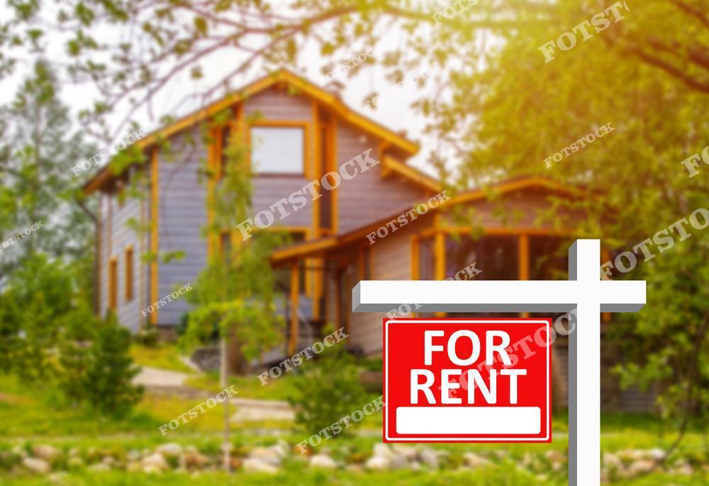 На переднем плане изображен знак "For Rent" красного цвета на белой стойке, установленный перед загородным деревянным домом, окруженным зеленью. Размытый фон подчеркивает естественную среду, создавая атмосферу уюта и спокойствия. Композиция символизирует доступность загородной недвижимости и комфортное жилье в природе.